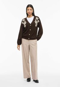 Sieviete, kas vilkti brūnu pogātu kardigānu ar beige ziedu izšuvumu, baltu kreklu, beige platām biksēm un melniem apaviem, stāv uz baltā fona.