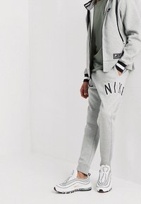 Šedá mikina na zip s černými akcenty, zelené tričko, šedé tepláky s logem "NIKE" a bílé sportovní boty s oranžovými detaily.