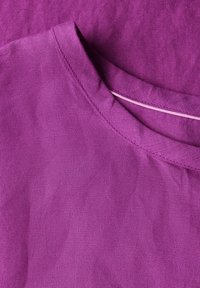 Camicia in tessuto viola con scollatura rotonda e una consistenza sottile. Dettaglio di cucitura visibile lungo il colletto e la cucitura della spalla.