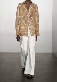 Tan linnen overhemd met een wit ruitpatroon, lange mouwen en knopen aan de voorkant, gecombineerd met witte wijde broek en zwarte sandaaltjes.