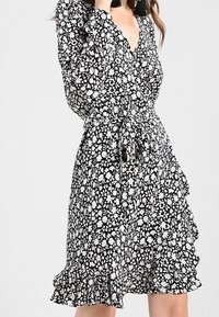 Vestido envolvente floral en blanco y negro con un escote en V profundo, mangas largas y un dobladillo con volantes. Presenta un cinturón de tela atado a la cintura.