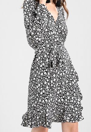 Robe portefeuille florale en noir et blanc avec un décolleté en V profond, des manches longues et un ourlet volanté. Elle possède une ceinture en tissu à la taille.