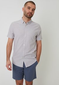 Camisa de botones de manga corta, color gris claro, con cuello, que presenta un sutil detalle de logo y un corte recto, combinada con pantalones cortos azul oscuro.
