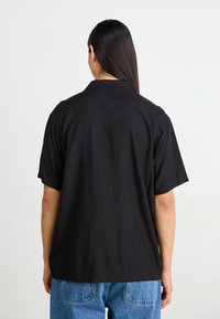 Chemise noire à manches courtes avec une coupe décontractée, dotée d'un col de chemise et d'une texture unie, portée sur un jean en denim bleu.