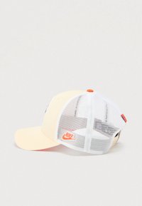 Béžová a bílá síťovaná kšiltovka Nike s oranžovým logem a knoflíkem, nastavitelným páskem vzadu, zobrazena z boku na bílém pozadí.