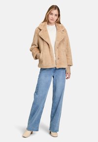 Giacca in pelliccia sintetica beige con ampi rever, due bottoni frontali e tasche laterali. Abbinata a jeans a gamba larga blu chiaro e scarpe beige.