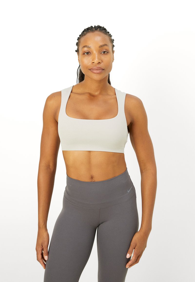 Lichtgrijze sportbeha met een vierkante halslijn en brede bandjes, gemaakt van rekbaar materiaal. Gepaarde met donkergrijze hoge taille legging.