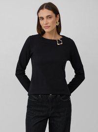Vrouw in een zwart blouse met lange mouwen en een gouden abstracte broche bij de halslijn, met een spijkerbroek aan, staand tegen een effen grijze achtergrond.