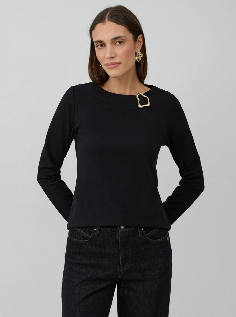 Vrouw in een zwart blouse met lange mouwen en een gouden abstracte broche bij de halslijn, met een spijkerbroek aan, staand tegen een effen grijze achtergrond.