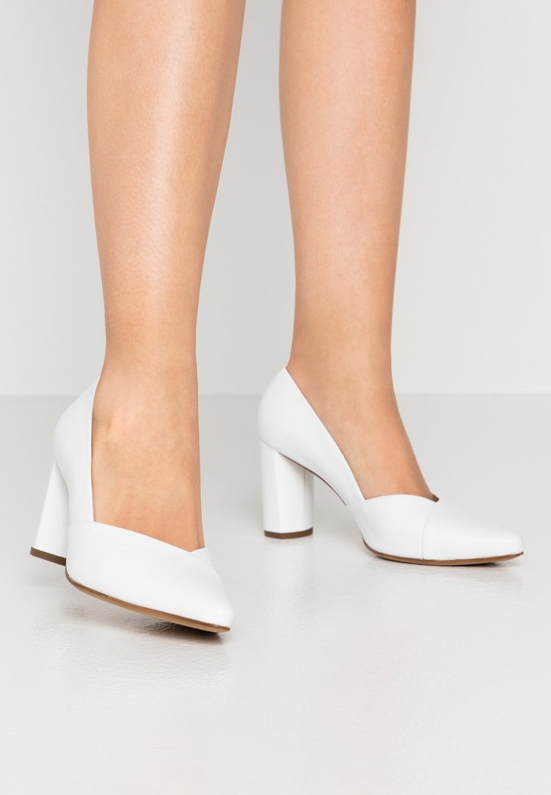 Femme portant des chaussures blanches à talons hauts avec des bouts arrondis, debout sur un sol lisse et clair.