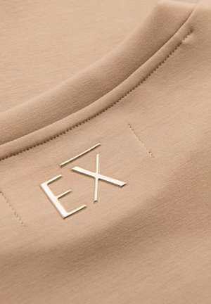 Beige stof med en syet søm og hævede metalbogstaver "EX" fastgjort nær sømmen.