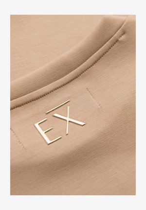 Béžová látka se šitým švem a vystouplými kovovými písmeny "EX" připevněnými poblíž švu.