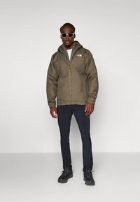 Olivzaļš jaka ar kapuci un rāvējslēdzēja aizdari, melnas bikses un melni pārgājienu zābaki. Modelis valkā saulesbrilles; stāv pret vienkāršu fonu.