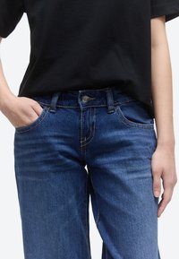 Άτομο που φορά σκούρο μπλε τζιν με το χέρι στην αριστερή τσέπη και ένα απλό μαύρο κοντομάνικο μπλουζάκι σε λευκό φόντο.