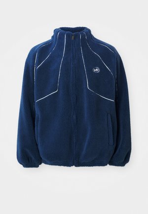 Navy blue fleece jacket with high collar, white piping design, zip closure, and side pockets. Soft texture with a relaxed fit.