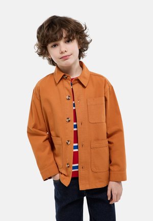 Enfant aux cheveux bouclés portant une veste orange boutonnée sur une chemise rayée rouge et blanche, avec une main dans la poche d'un pantalon sombre.