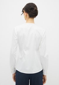 Blusa bianca a maniche lunghe con una texture liscia, orlo arrotondato e polsini con bottoni. La vista posteriore mostra una vestibilità sagomata e una leggera lucentezza.