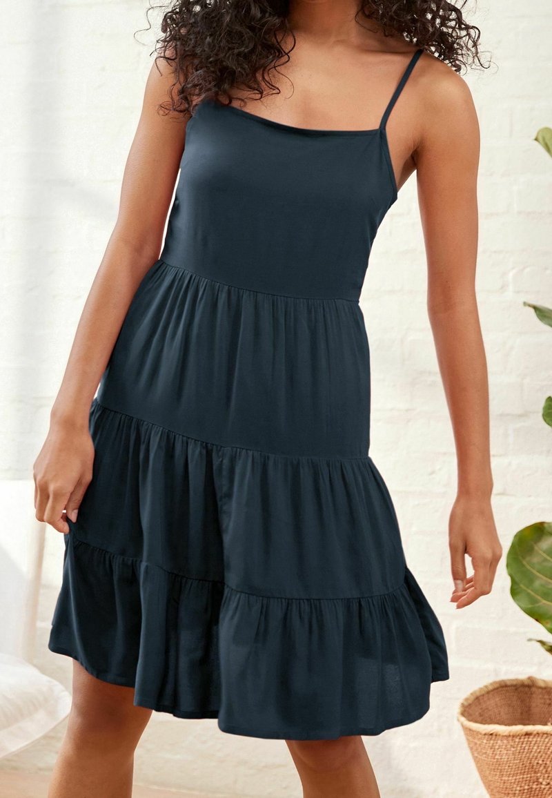 Femme portant une robe midi noire sans manches à volants avec de fines bretelles, debout à l'intérieur près d'une plante et d'un panier.