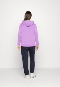 Felpa viola con cucitura centrale sul retro e cappuccio regolabile, abbinata a pantaloni della tuta blu navy e sneakers bianche con dettagli rossi.