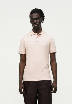 Νέος άνδρας με σκούρο δέρμα και πλεξούδες, φορώντας ένα ανοιχτό ροζ κοντομάνικο πόλο και σκοτεινό παντελόνι, στέκεται μπροστά σε ένα απλό φόντο.