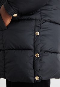 Close-up van een zwart doorgestikt jack met gouden drukknopen en een hand deels zichtbaar in de zak.