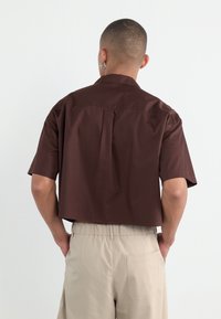 Brown short-sleeve shirt with a straight cut, featuring a back pleat. Worn with light beige elastic waist trousers, showing a pocket detail.