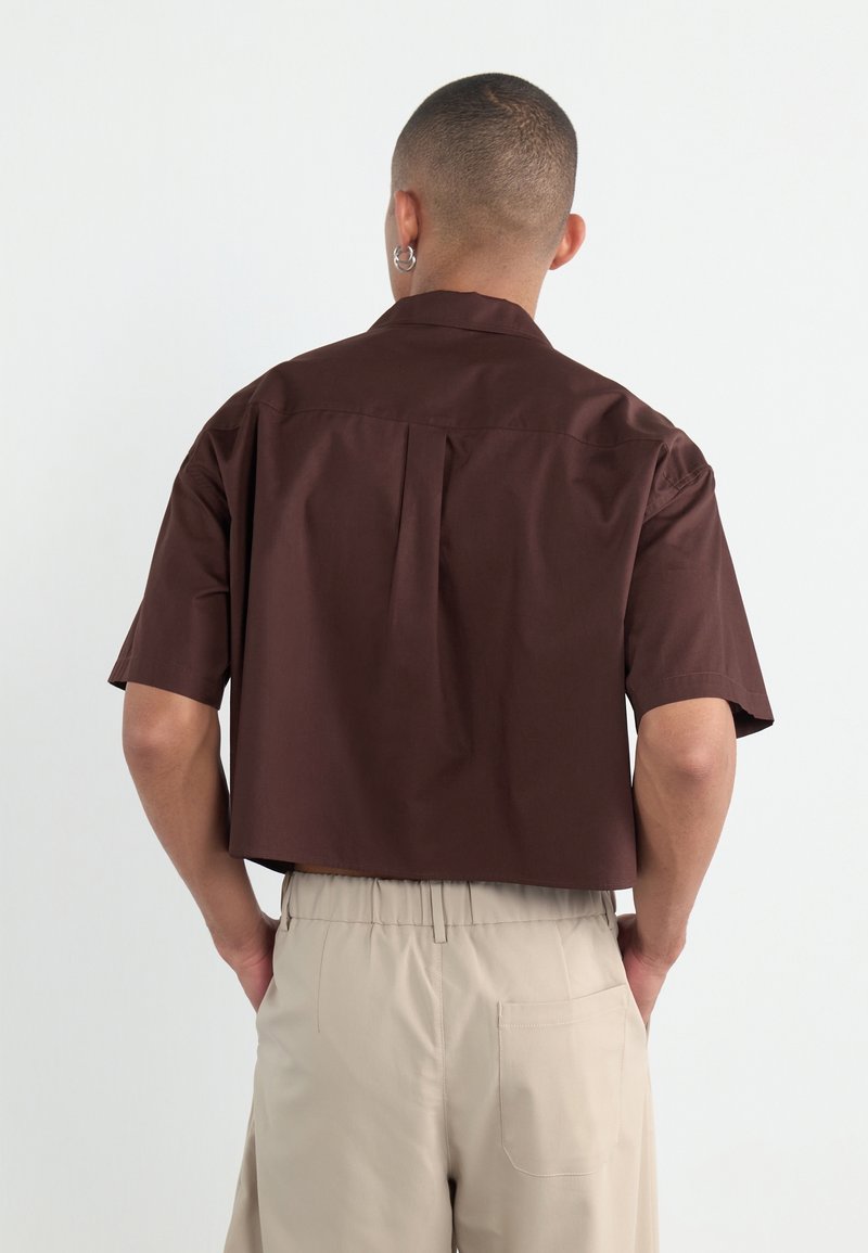 Brown short-sleeve shirt with a straight cut, featuring a back pleat. Worn with light beige elastic waist trousers, showing a pocket detail.