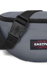 Marsupio in tessuto grigio con una tracolla nera, dotato di una fibbia in plastica e di una patta nera con logo e lettering rosso. Chiusura con zip sulla parte superiore.