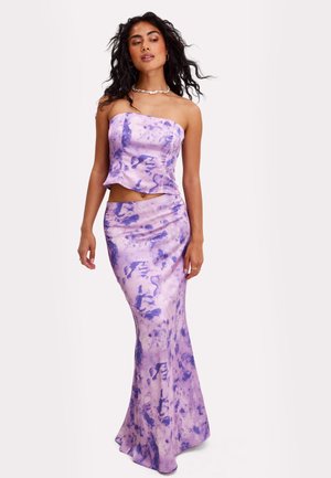 Femme portant un haut sans manches et sans bretelles tie-dye lilas et violet ainsi qu'une jupe longue assortie, debout devant un fond uni.