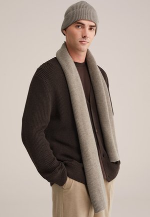 Brauner gerippter Strickcardigan über einem dunklen Hemd, kombiniert mit einem hellgrauen gerippten Schal und passender Mütze, vor einem neutralen Hintergrund.