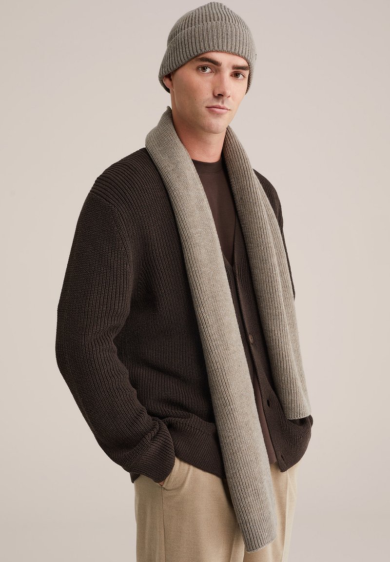 Browninga ribota trikotāžas kardigans virs tumša krekla, apvienots ar gaiši pelēku ribotu šalli un atbilstošu cepuri, uz neitrāla fona.