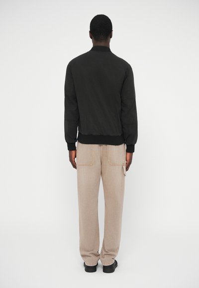 Μαύρο μπουφάν bomber με ριμπ μανσέτες και τελείωμα, συνδυασμένο με μπεζ παντελόνι φαρδιάς γραμμής που διαθέτει πίσω τσέπες και εμφανείς ραφές.