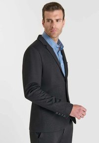 Blazer su misura grigio scuro con una texture liscia, rever a lesina e dettagli a cinque bottoni sui polsini, indossato sopra una camicia colletto azzurra.