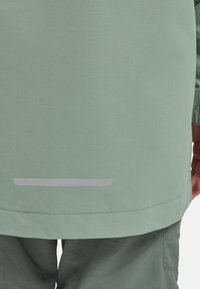 Bas du dos d'une personne portant une veste vert clair avec une bande réfléchissante horizontale au-dessus de l'ourlet et un pantalon assorti.