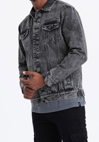 Homme portant une veste en jean noire délavée sur une chemise grise et un pantalon cargo noir, ajustant sa manche gauche avec les deux mains.