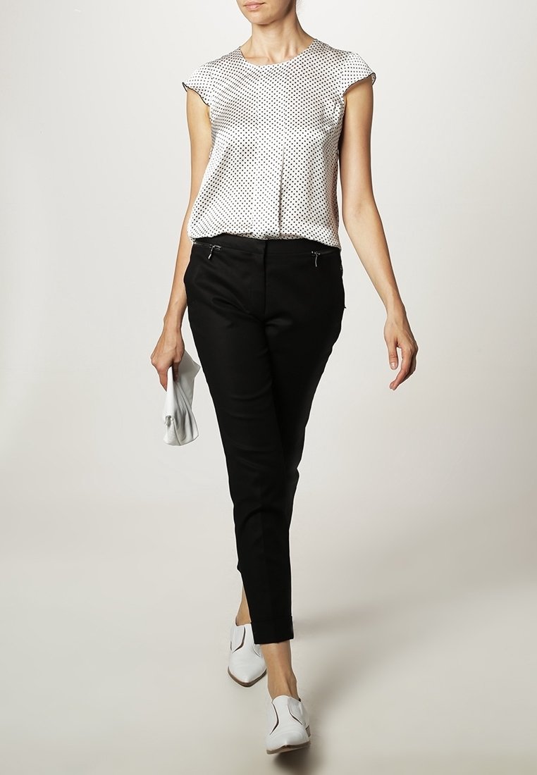 Femme marchant, portant un chemisier blanc à pois, un pantalon noir, des chaussures blanches, et tenant une petite pochette blanche sur un fond uni.