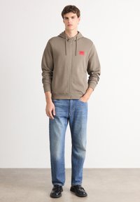 Sudadera verde oliva con cremallera y cordón, con un parche de logo rojo, combinada con jeans rectos azul claro y zapatos negros.