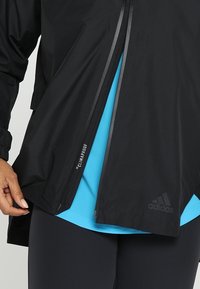 Personne portant une veste Adidas noire avec fermeture éclair partiellement ouverte, révélant une chemise bleue en dessous et une étiquette "Climaproof" sur la fermeture éclair.