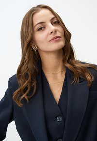 Blazer navy con due bottoni, indossato su un top a scollatura a V profonda. Collana a catena dorata e orecchini geometrici dorati coordinati.
