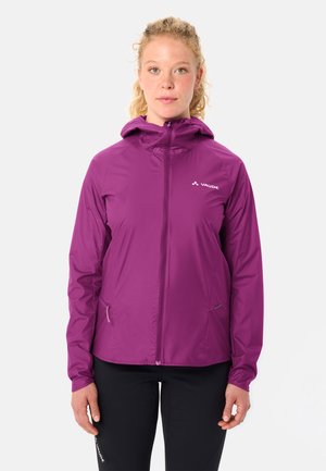 Frau trägt eine lila Kapuzenjacke mit Reißverschluss und schwarze Hose, steht vor einem schlichten weißen Hintergrund.