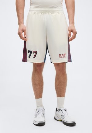 Férfi krémszínű atlétikai rövidnadrágot visel piros és fekete csíkokkal az oldalán, elöl a 77-es szám és EA7 Emporio Armani logók láthatók.