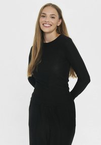 Woman with long light brown hair smiling, wearing a black long-sleeve top and black pants, standing against a plain light background.
