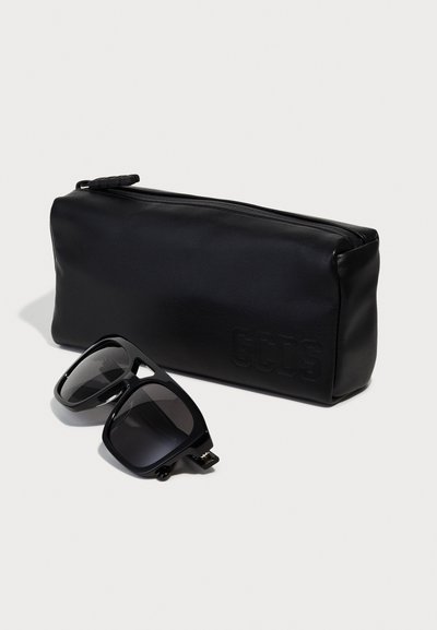 Estuche negro de cuero con cremallera y logotipo "GCDS" en relieve, junto a unas gafas de sol negras de gran tamaño con acabado brillante y lentes oscuros.