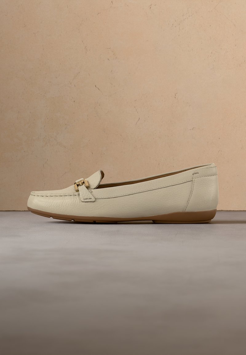 Mocassin en cuir beige avec un petit détail de boucle dorée et une semelle en caoutchouc marron, présenté sur un fond texturé neutre.