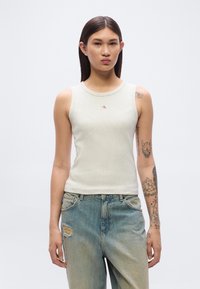 Ανοιχτό γκρι αμάνικο τοπ με στρογγυλή λαιμόκοψη, διακοσμημένο με ένα μικρό κόκκινο λογότυπο. Συνδυάζεται με τζιν παντελόνι μπλε χρώματος με φθορές. Λείο βαμβακερό ύφασμα.