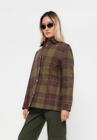 Rutig skjorta i olivgrön, vinröd och svart; knappstängd design med krage och en enskild bröstficka; bärs med mörkgröna byxor.