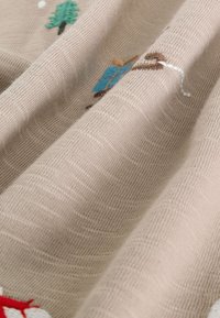 Tissu beige avec un tissage texturé, comportant des éléments brodés colorés comme des arbres et un accent rouge, créant un design ludique.