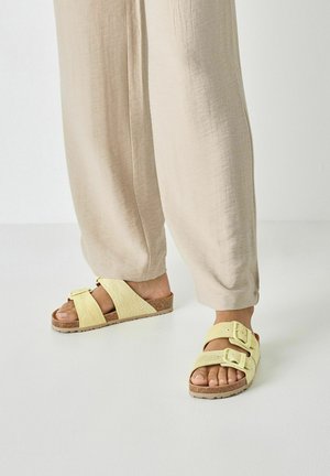Gele sandalen met een kurken zool, voorzien van twee brede riemen met gespsluitingen en een bladmotief, gecombineerd met beige broek.