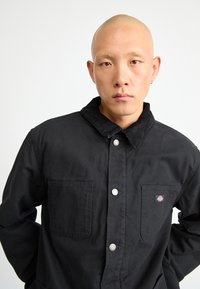Bald man with small hoop earring wearing black button-up jacket with two chest pockets, one displaying a small colorful logo.
