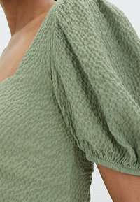 Groene textuurtop met een diepe halslijn en pofmouwen. De stof heeft een gekreukt patroon en elastische manchetten voor de juiste vorm.